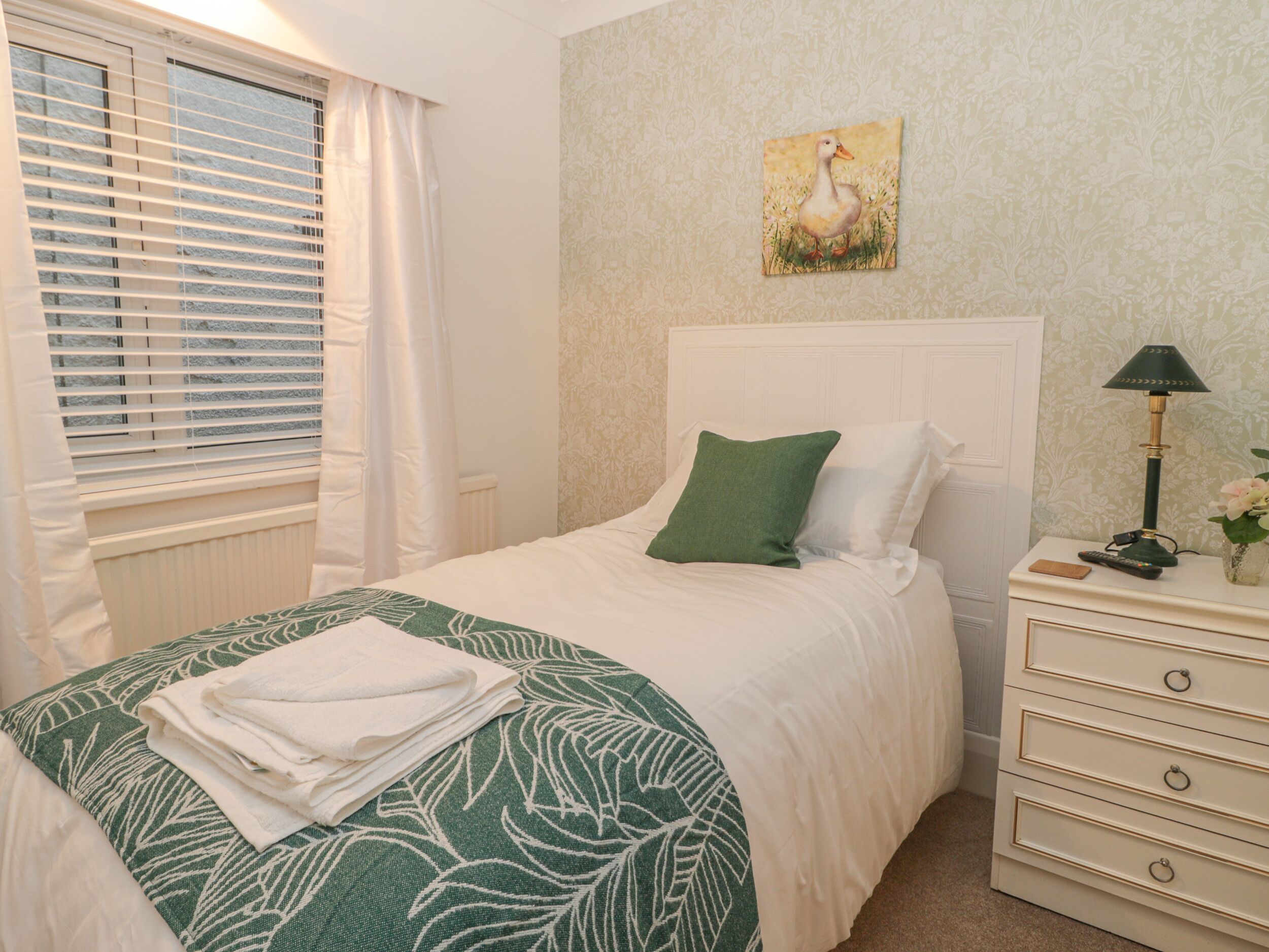 A neatly made bed with a green leaf-patterned bedspread and white pillows, a bedside table with a lamp and remote controls, and a window with blinds and a curtain.