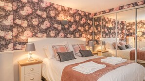 A bedroom with a floral wallpaper, a large bed with multiple pillows, a nightstand with a lamp, and a mirror on the wall. - Hillcrest (Scarborough)