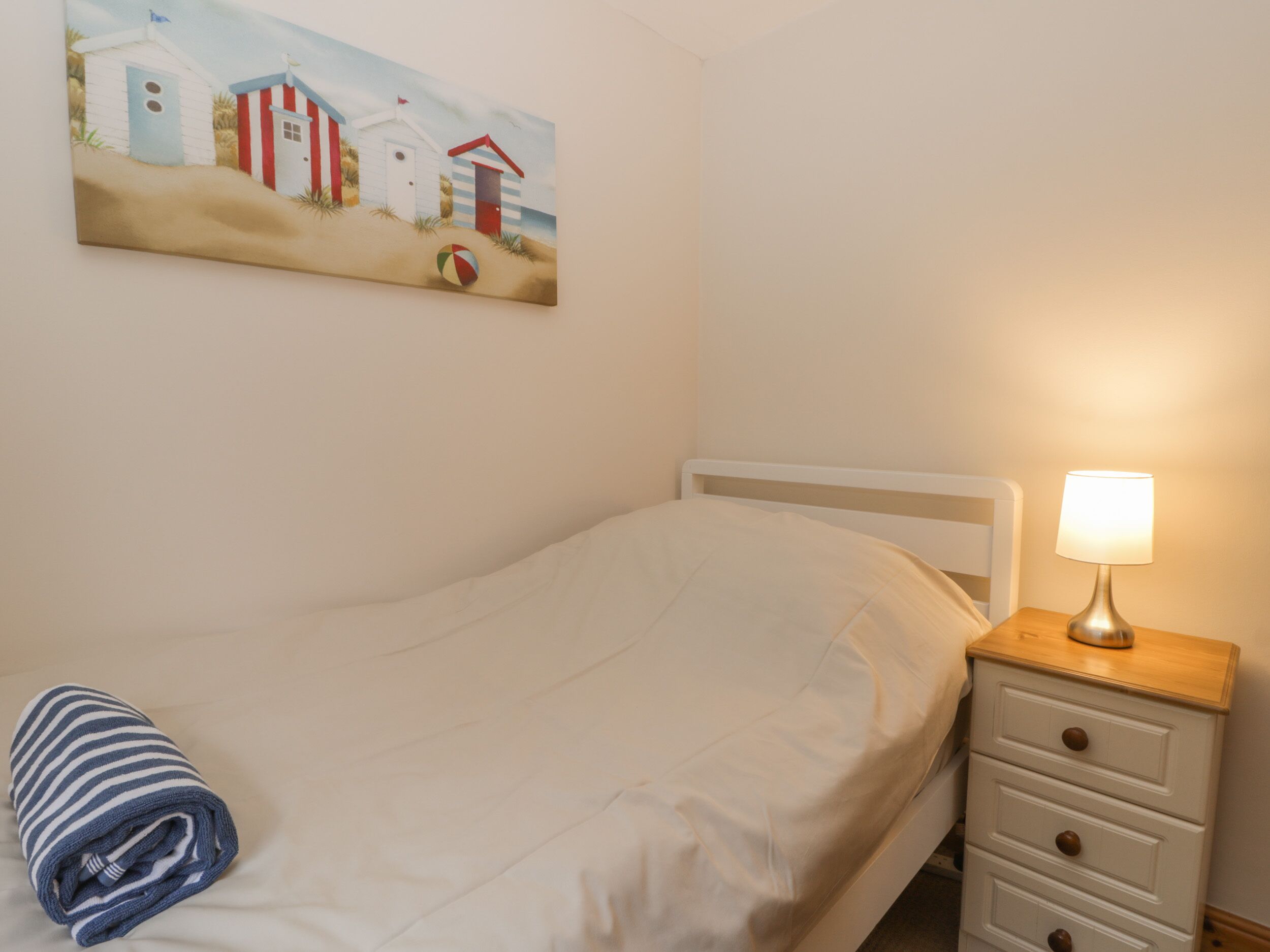 A bedroom with a bed, a nightstand, a lamp, a rolled towel, and a beach-themed painting on the wall.