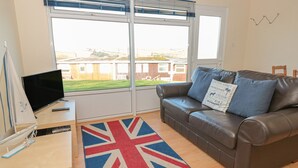 Ett vardagsrum med en soffa i läder, en matta med Union Jack-mönster, en tv på ett träbord och stora fönster med persienner.