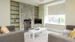 A living room with a stone fireplace, a flat-screen TV, and a large window with a view of the outside. - Maes-yr-haf (Criccieth)