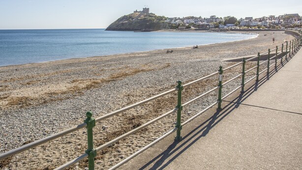 A coastal promenade with a pebble beach, a castle-like structure on a hill, and a row of houses in the background. - Maes-yr-haf (Criccieth)