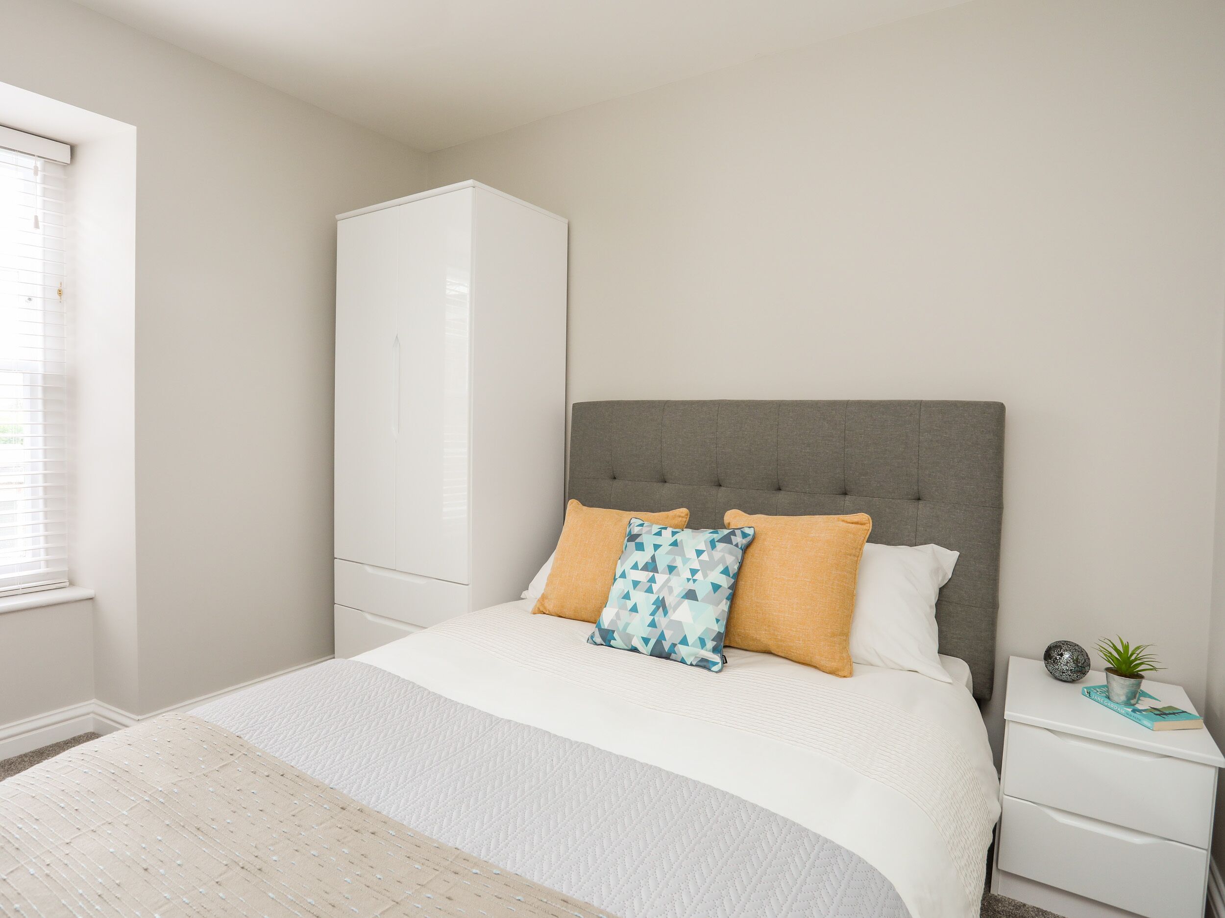 Une chambre à coucher avec un lit, une tête de lit rembourrée de couleur grise, du linge de lit blanc, une armoire blanche, une table de chevet avec une plante et une fenêtre avec des stores.