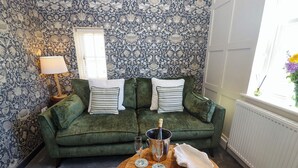 A green sofa with striped pillows, a wooden table with a wine glass and bottle, and a patterned wallpaper. - Gavel Cottage (Whitby)