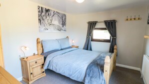 A bedroom with a bed, wooden nightstands, a desk, a window with curtains, a painting on the wall, and a ceiling light. - Castle View Cabin (Scarborough)