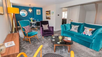 A modern living room with teal and purple furniture, a glass coffee table, and a dining area in the background.