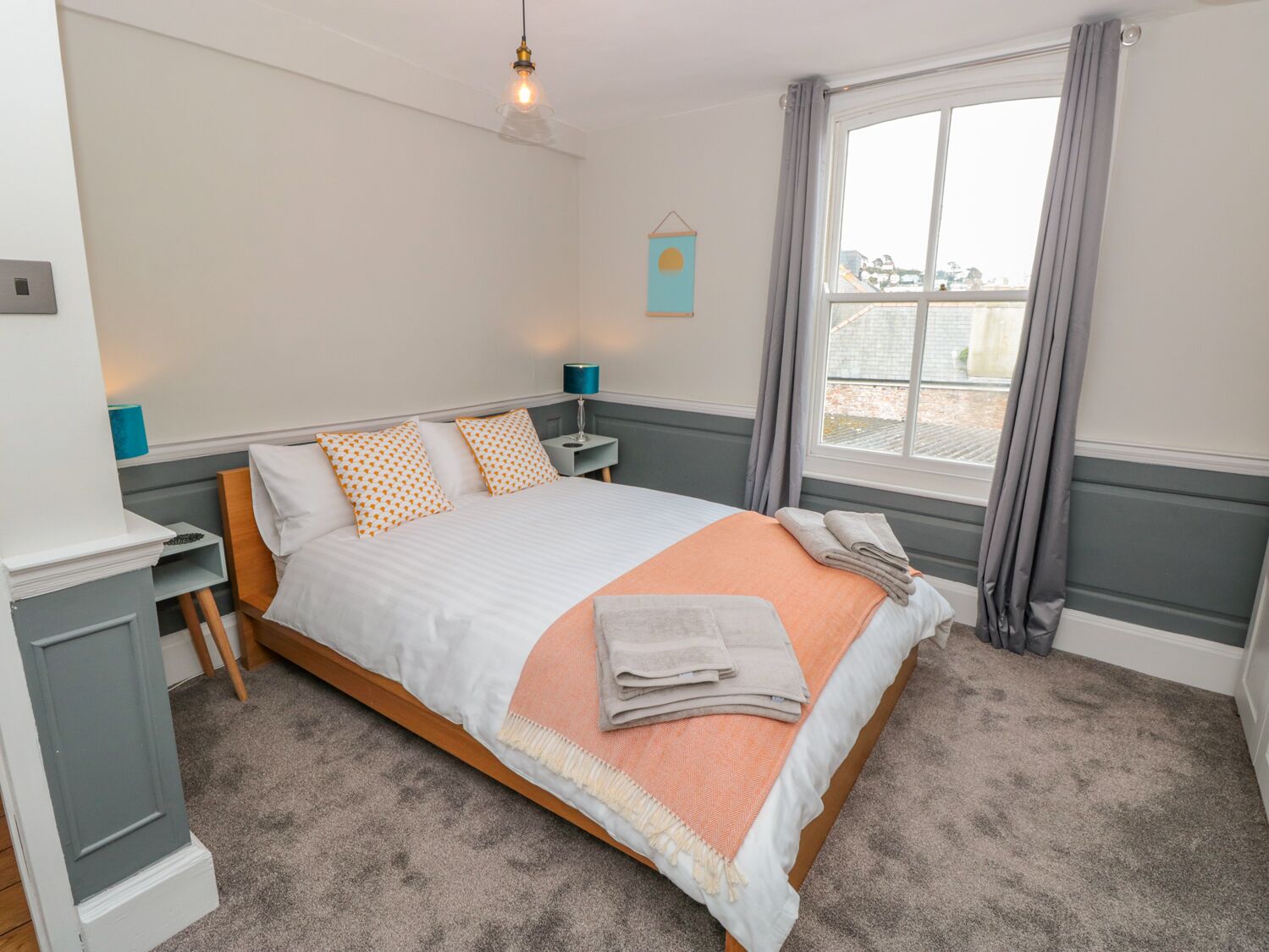 Un dormitorio ordenado con cama, mesitas de noche, lámpara y ventana con cortinas.