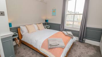 Un dormitorio ordenado con cama, mesitas de noche, lámpara y ventana con cortinas.