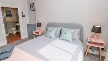Un dormitorio con una cama, dos mesitas de noche, una mesita auxiliar, un cuadro en la pared y un baño visible a través de una puerta abierta.