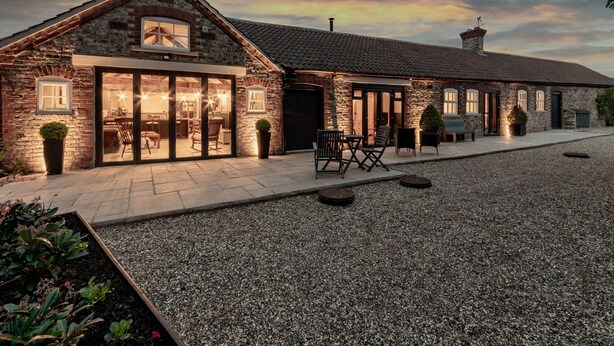 A stone building with a large glass door entrance, a patio with outdoor seating, and a gravel driveway. - Sykes Lodge (Malton)
