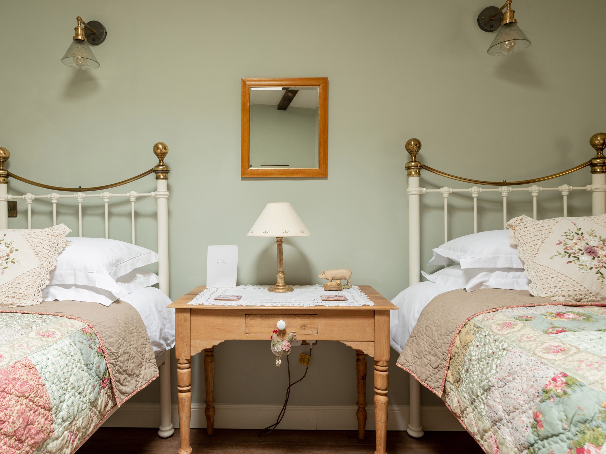 A room with two white metal beds, a wooden nightstand with a lamp, a mirror, and a small decorative item on the nightstand.