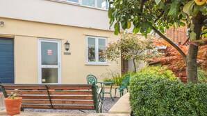 Un banco frente a un edificio con puerta y ventanas, un pequeño jardín con seto y un árbol con hojas otoñales.