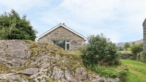 Une maison en pierre avec un toit en pente, entourée d’une pelouse et d’une colline rocheuse.