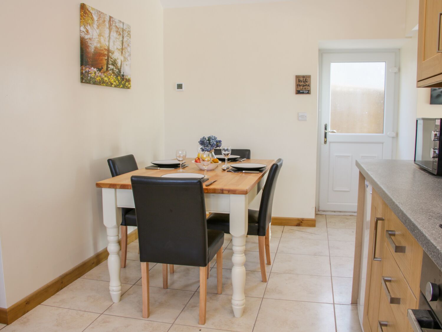 Een eetgedeelte met een houten tafel voor vier personen, omringd door zwarte stoelen. Aan de muur hangt een schilderij van bomen en bloemen.