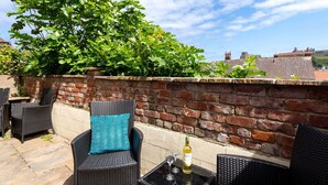 Eine Terrasse im Freien mit Korbmöbeln, einer Backsteinmauer und Blick auf Gebäude im Hintergrund.
