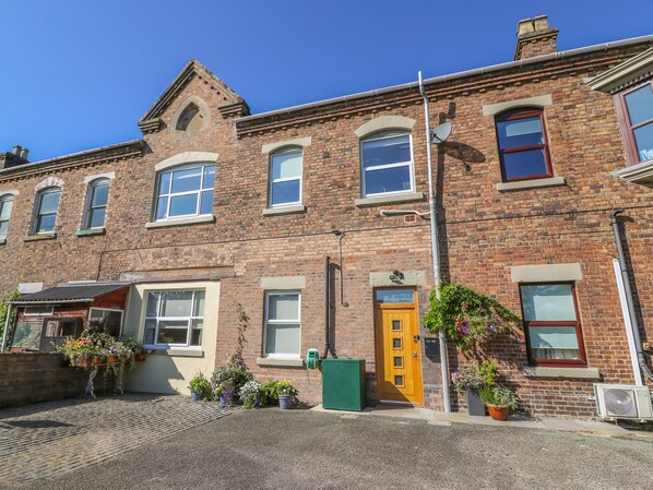 A two-story brick building with a traditional design, featuring a central entrance with a wooden door, multiple windows, and a small garden area with potted plants.