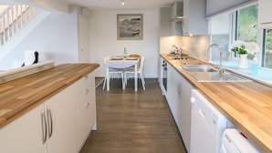A compact kitchen with wooden countertops, white cabinetry, a built-in dishwasher, and a dining area with a table and chairs. - Cuckoo Cottage (Bridport)