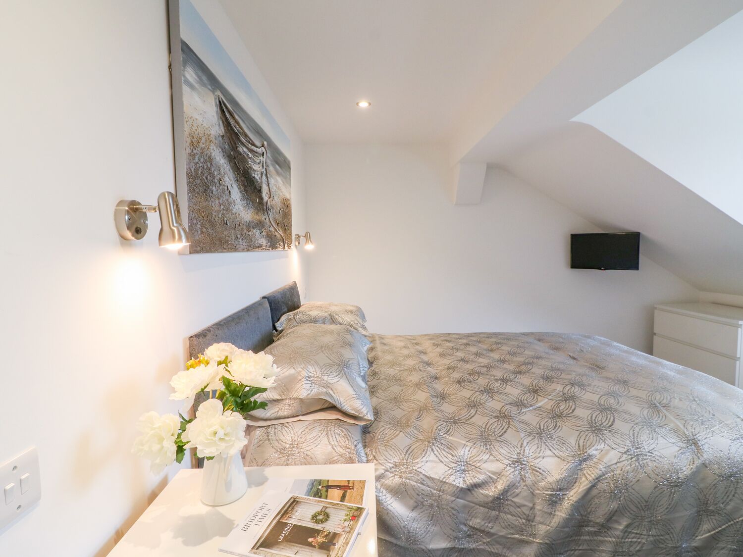 A bedroom with a bed, a bedside table with a vase of flowers, a painting on the wall, and a television on the ceiling.