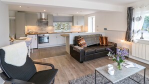 A modern living room with a sofa, a coffee table, and a dining area with a kitchen in the background. - Cuckoo Cottage (Bridport)