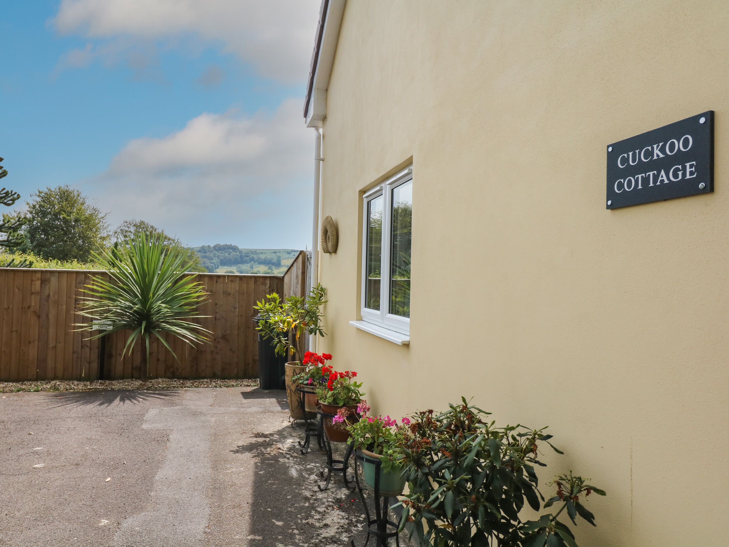 A cottage named 'CUCKOO COTTAGE' with a wooden fence, potted plants, and a view of the countryside.
