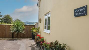 A cottage named 'CUCKOO COTTAGE' with a wooden fence, potted plants, and a view of the countryside.