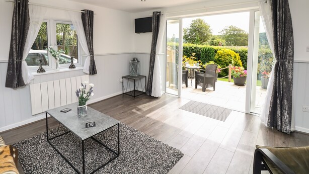 A room with a dining table, chairs, a small side table with a vase, a radiator, and a view of a garden through a glass door. - Cuckoo Cottage (Bridport)