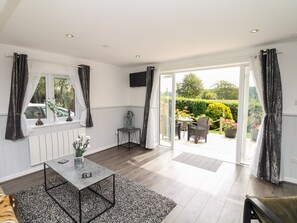 A room with a dining table, chairs, a small side table with a vase, a radiator, and a view of a garden through a glass door. - Cuckoo Cottage (Bridport)
