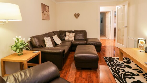 A living room with a sectional sofa, ottoman, wooden coffee table, and a small side table with a lamp. There is a heart-shaped decoration on the wall and a plant on the side table. - 7 Riverside Gardens (Inverness)