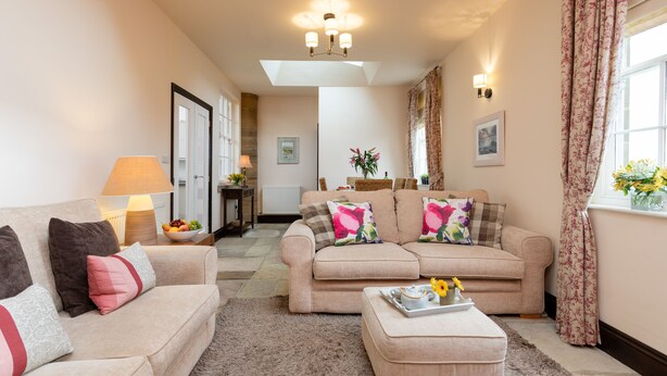 A living room with a beige sofa, a matching ottoman, and a lamp. There is a window with curtains and a vase of flowers on the windowsill. - Mews Cottage (Whitby)