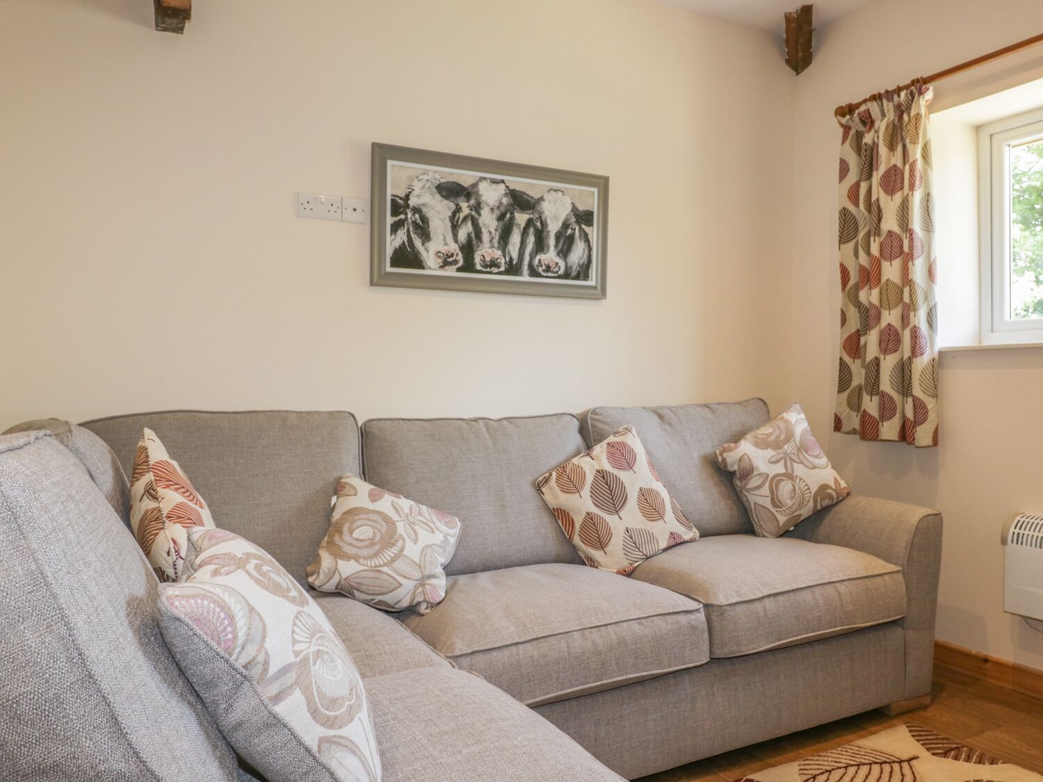 Un soggiorno con un divano grigio, cuscini decorati, un quadro con bovini e una finestra con tende a motivi fogliacei.