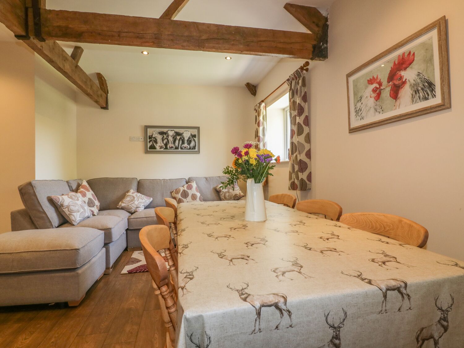 A cozy living room with a sofa, a dining table with a deer-patterned tablecloth, and a painting of chickens on the wall.