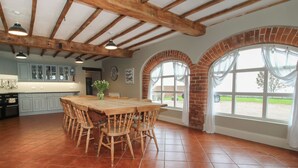 Une cuisine spacieuse avec une grande table et des chaises en bois, une cheminée en briques et de grandes fenêtres avec des rideaux.
