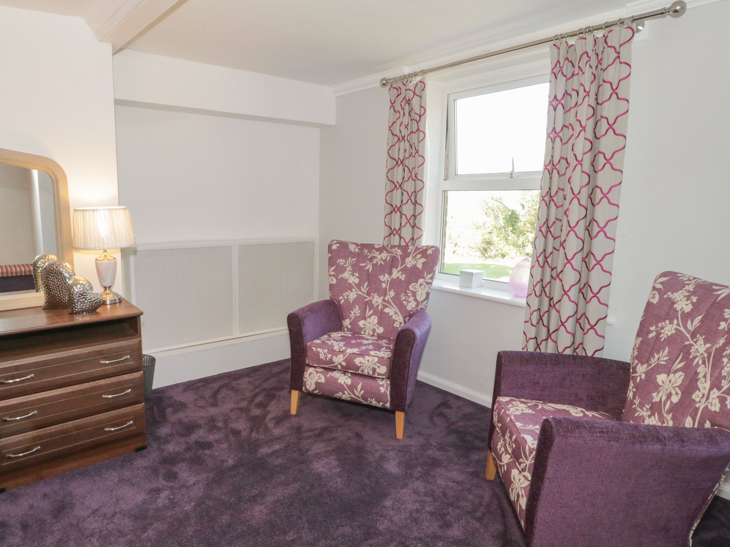 A room with a purple carpet, two purple armchairs with floral upholstery, a wooden dresser with drawers, a mirror, and a window with patterned curtains.