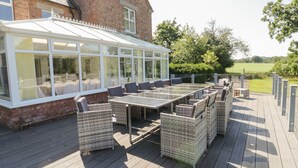 A conservatory with glass walls, a wooden deck, wicker furniture, and a brick building in the background. - Maes Elwy (Saint Asaph)