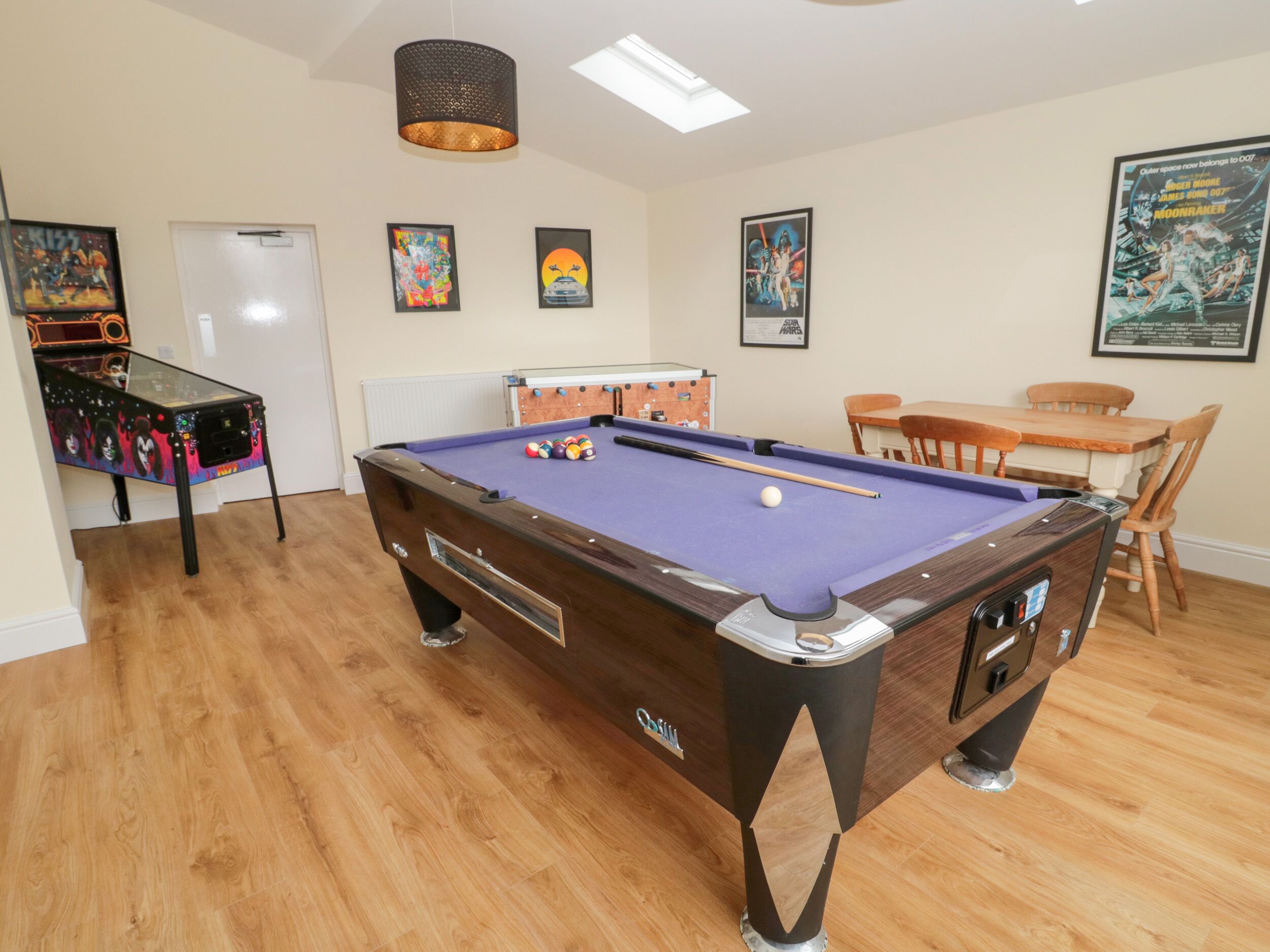 A pool table with a blue felt surface, a pool cue, and several balls. There is a wooden table with chairs in the background, and a ceiling light fixture above.