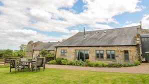 A stone building with a sloped roof, large windows, and a grassy lawn with outdoor furniture. - The Steadings (Morpeth)