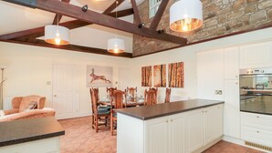 A spacious kitchen with a central island, white cabinetry, and a stone wall. There are hanging pendant lights and a dining area with wooden chairs. - The Steadings (Morpeth)