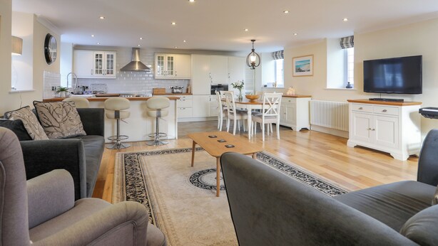 A modern living room with a sofa, a coffee table, and a TV. There is a kitchen area with white cabinets and a dining table with chairs. - Capel Hermon (Holyhead)