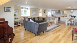 A spacious living area with a grey sofa, wooden flooring, a dining table with chairs, and a kitchen in the background. - Capel Hermon (Holyhead)