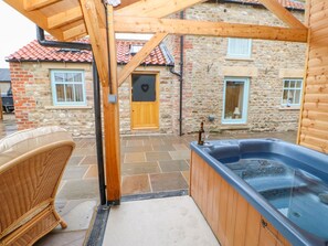 A wooden hot tub in a covered outdoor area with stone flooring, a wicker chair, and a stone building with a red-tiled roof in the background. - The Dove Cote (Richmond)