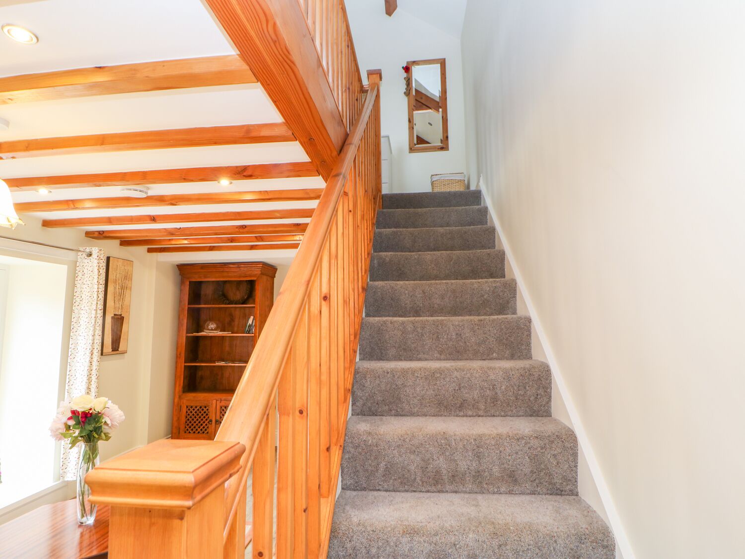 Un escalier avec des rampes en bois et des marches recouvertes de moquette, menant à un palier où se trouvent un meuble en bois et un vase de fleurs.