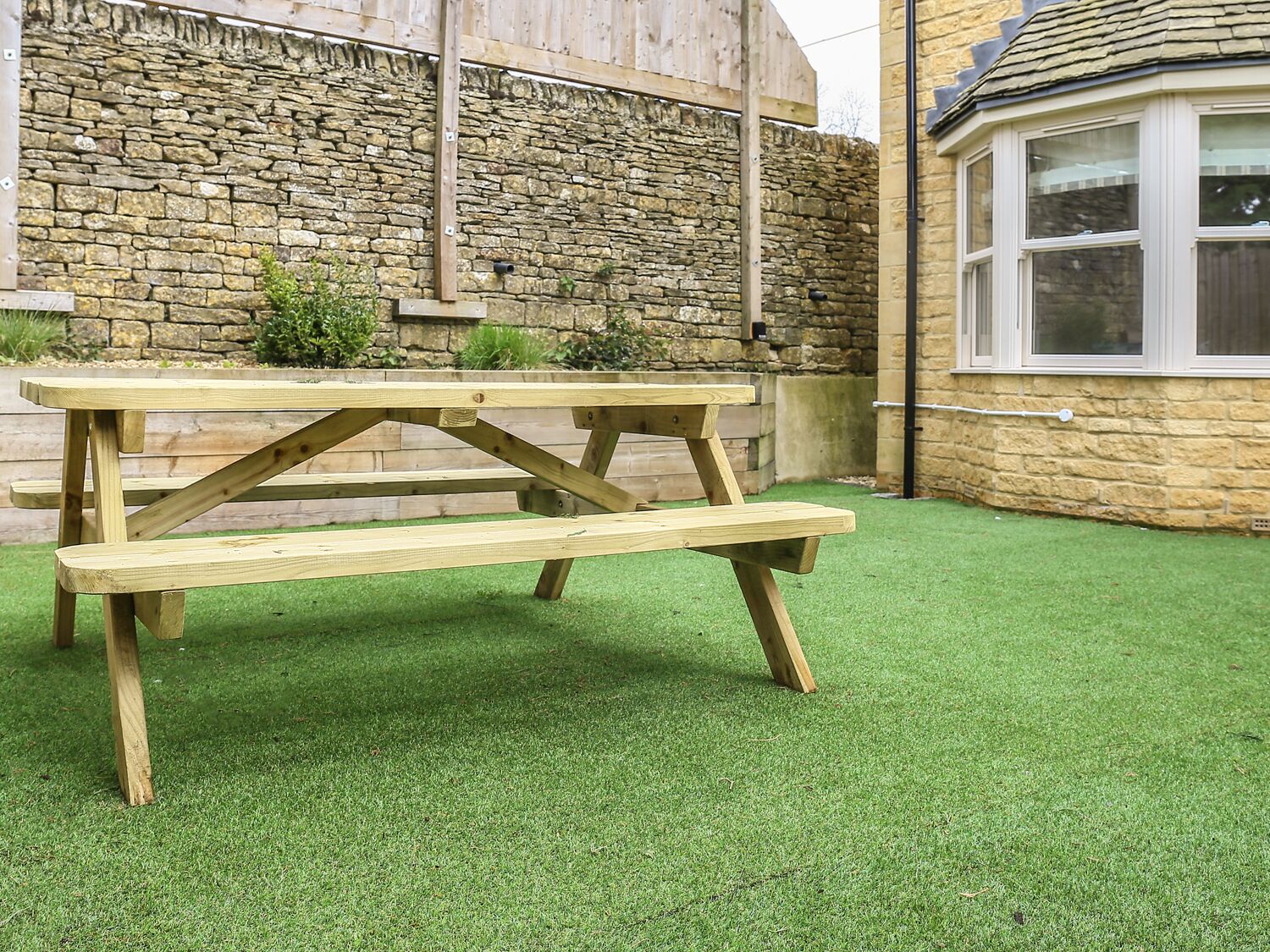 A wooden picnic table in a garden with a stone wall and a window in the background.