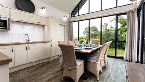 A modern kitchen with white cabinets, a dining table set for four, and a large window offering a view of greenery. - Betony (Bamburgh)