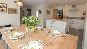 A dining area with a wooden table set for six, white cabinets, and a kitchen in the background. - Willow Cottage (Dartmouth)