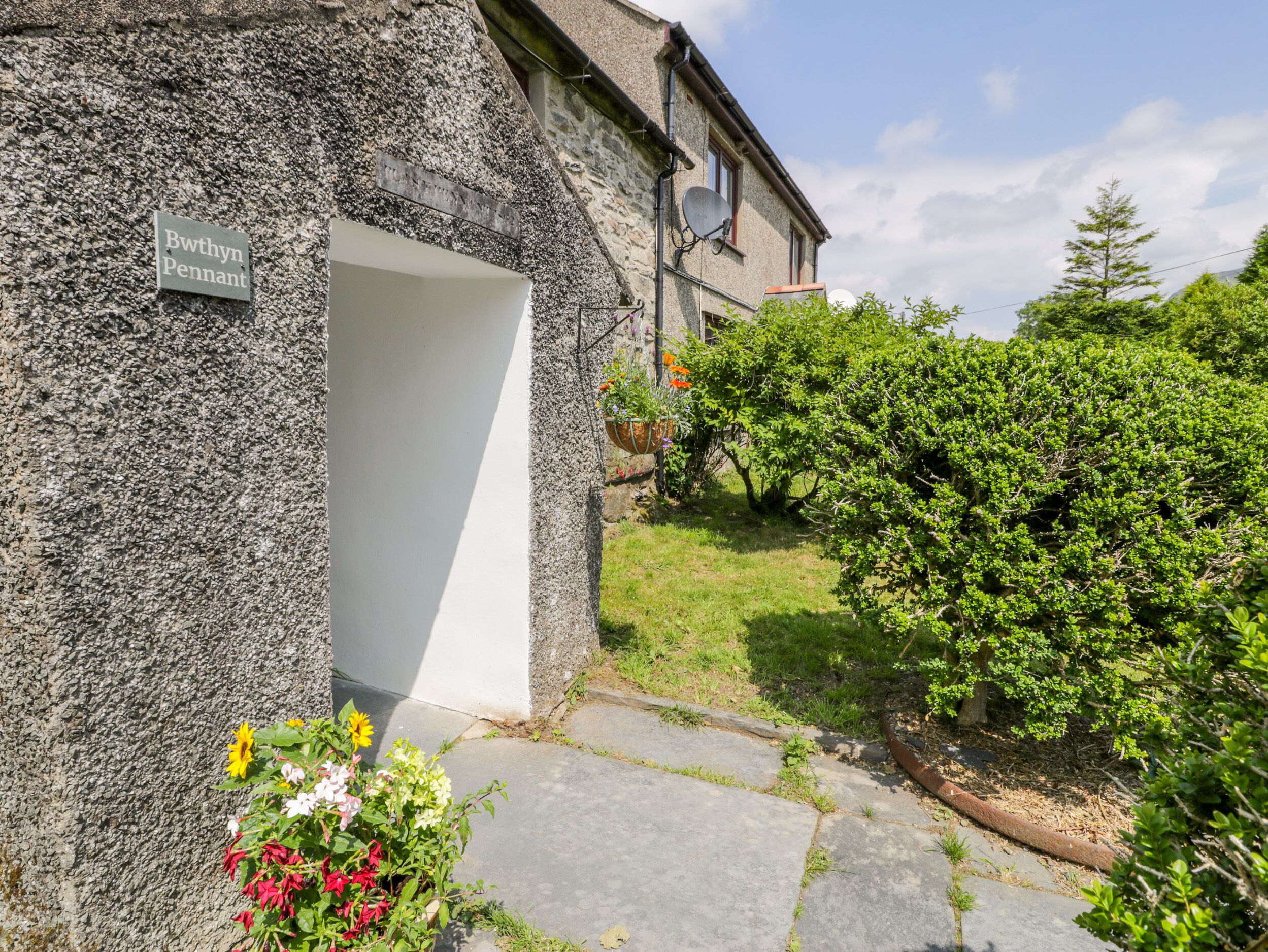 A stone building with a white door, a sign reading "Bwithyn Pennant", and a garden with various plants and flowers.