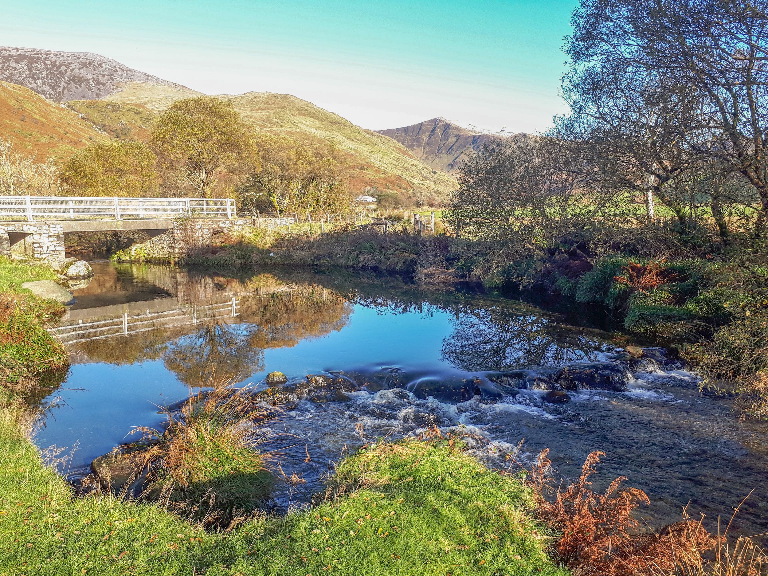 A river with a stone bridge, surrounded by lush greenery and hills.
