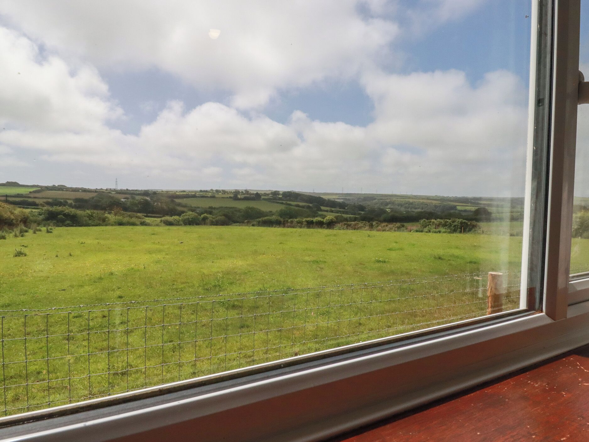 Vista a través de una ventana: un campo verde con una cerca, cielo despejado y colinas al lejano.