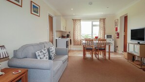 A living room with a sofa, a dining table with chairs, a kitchen area, a television, and a fire extinguisher. - Little Lanes (Camelford)