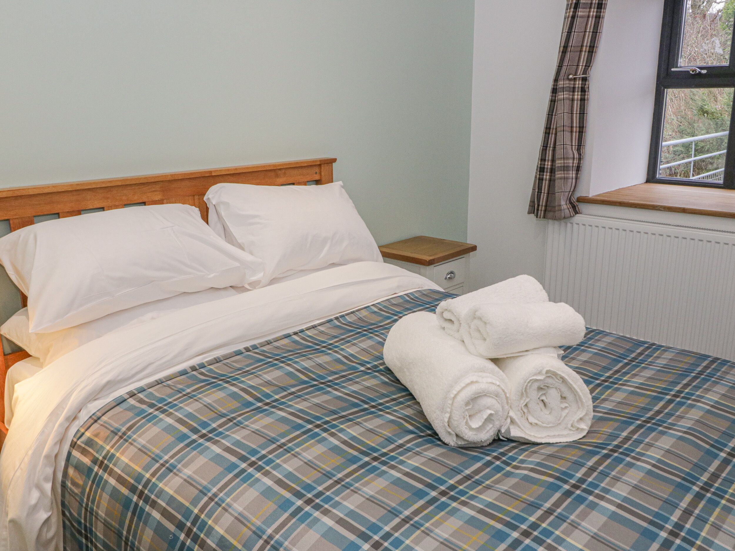 A neatly made bed with white linens and a plaid bedspread, a wooden nightstand with a drawer, and a window with a view of greenery.