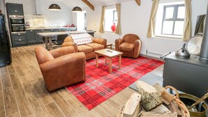 A spacious living area with a sofa set, a coffee table, and a red plaid rug. There is a fireplace with a kettle on top, and a kitchen area with modern appliances and a window with curtains.